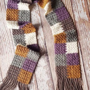 May include: A crocheted scarf with a checkered pattern in shades of purple, brown, white, and mustard yellow. The scarf has long brown fringe on both ends.