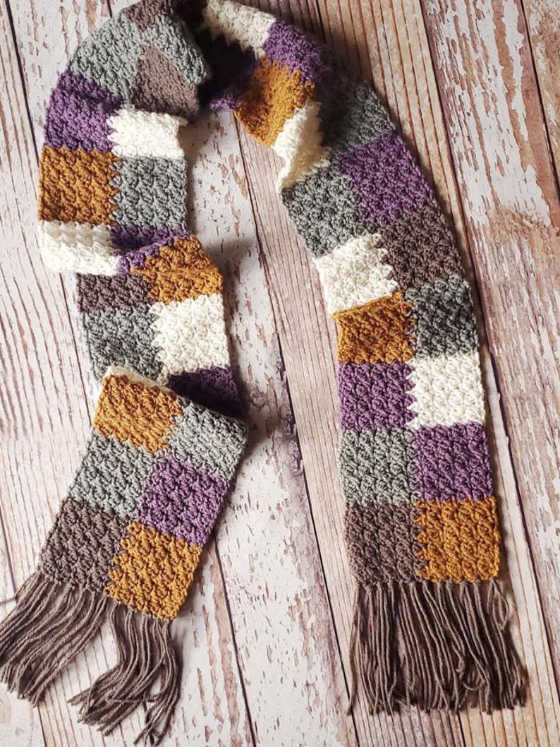 May include: A crocheted scarf with a checkered pattern in shades of purple, brown, white, and mustard yellow. The scarf has long brown fringe on both ends.