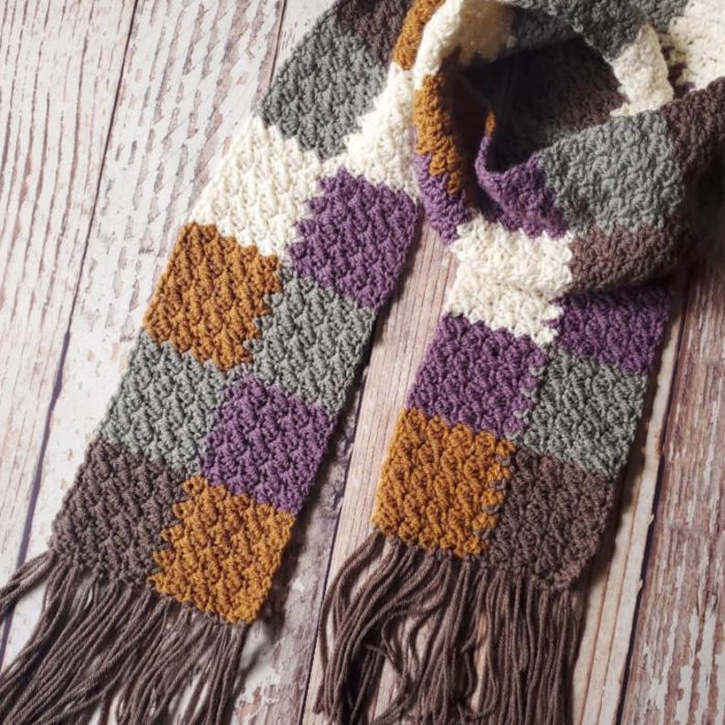 May include: A crocheted scarf with a geometric pattern in shades of brown, purple, grey, and white. The scarf has long fringe on both ends.