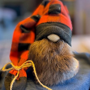 May include: A brown gnome with a long, bushy beard wearing a red and black plaid hat. The hat is tied with a brown twine bow.