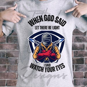 Può includere: T-shirt grigia con grafica di un saldatore e la scritta "When God Said Let There Be Light I Said Watch Your Eyes". La grafica del saldatore è a forma di scudo con una combinazione di colori blu e giallo.
