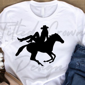 Pode incluir: Silhueta preta de uma mulher a cavalo. A mulher está usando um chapéu de cowboy e botas. O cavalo está correndo. A imagem está em uma camiseta branca.