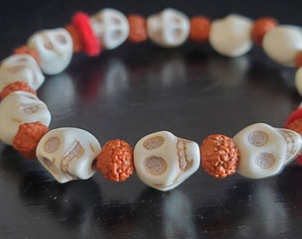 May include: A white skull bead bracelet with orange beads. The bracelet has a red string accent.