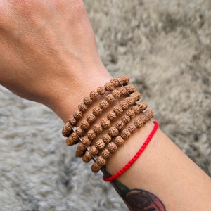 May include: A stack of five brown beaded bracelets with a red string bracelet on the wrist. The bracelets are made of small, round beads.
