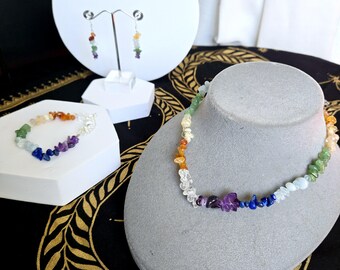 May include: A rainbow gemstone necklace, bracelet, and earrings set. The necklace is made with small, colorful stones and is displayed on a gray mannequin. The bracelet and earrings are displayed on a white stand.