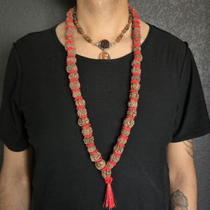 May include: A long brown mala necklace with red string accents and a tassel. The necklace is worn by a person and has a smaller silver necklace with a pendant underneath.
