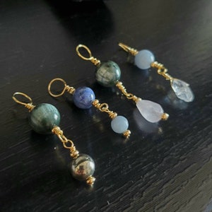 May include: Four gold-wrapped gemstone charms with different colored stones. The charms feature a green stone, a blue stone, a light blue stone, and a pink stone.