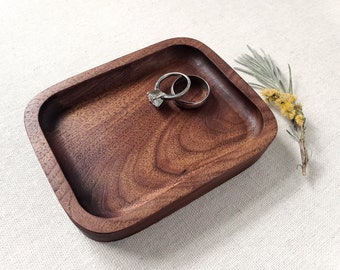 May include: A small, rectangular wooden tray made of dark walnut wood. The tray has rounded edges and a smooth, polished finish. Two silver rings are resting in the center of the tray.