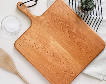 May include: A square wooden cutting board with a handle and a brown leather hanging loop. The board features a prominent natural wood grain pattern. The text NEW SIZES! is printed in white at the bottom of the image against a gray background.