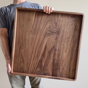 May include: A large square wooden serving tray crafted from dark brown walnut wood with a visible natural grain pattern. The tray features a flat base and raised edges with rounded corners, providing a smooth finish and a sturdy construction.