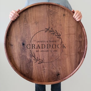 May include: A large, round wooden serving tray with a dark brown stain. The tray has a personalized inscription that reads "Jayson & Tanya Craddock Est. January 18, 1994" with a decorative wreath design.
