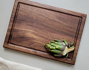 May include: A large, rectangular walnut cutting board with a dark brown finish and a raised edge. An artichoke is resting on the board.