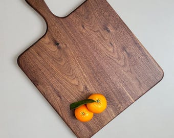 May include: A square dark brown wooden serving board with a handle and a leather loop at the top. The board features a prominent wood grain pattern and is positioned with two bright orange tangerines with a green leaf resting on its surface.