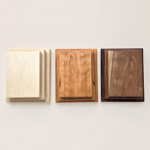 May include: Three rectangular wooden display stands arranged side by side. The left stand is light cream-colored wood, the center stand is medium-toned amber wood, and the right stand is dark brown wood. Each features a raised, centered platform.