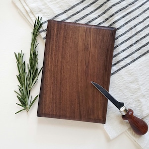 May include: A rectangular, dark brown wooden cutting board with a small cheese knife. A sprig of rosemary and a white and black striped cloth are also present. The cutting board is likely used for food preparation and serving.