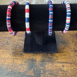 May include: Three bracelets made of red, white, and blue beads. The bracelets are stacked on a black display stand.