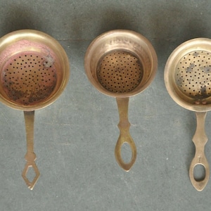 May include: Three antique brass tea strainers with handles. Each strainer has a round, perforated bowl with a small, ornate handle.