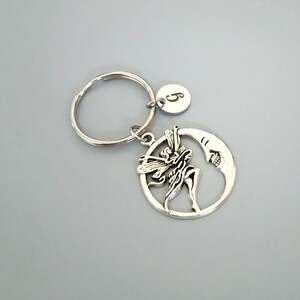 Könnte beinhalten: Silberner Schlüsselanhänger mit einem Anhänger einer Fee, die um einen Halbmond fliegt. Der Anhänger hat eine kleine silberne Scheibe mit dem eingravierten Buchstaben "S".