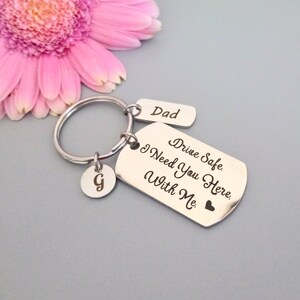 May include: Silver keyring with three tags. The largest tag reads "Drive Safe. I Need You Here. With Me." and has a small heart at the bottom. The middle tag reads "Dad". The smallest tag is round and has the letter "G" on it.