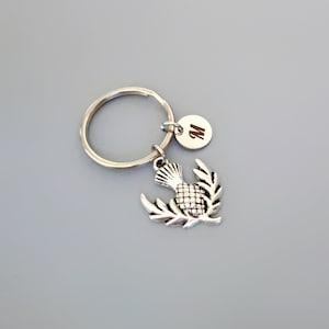 Pu&ograve; includere: Portachiavi d'argento con un ciondolo a forma di cardo d'argento e un ciondolo a disco d'argento con la lettera "M" incisa.