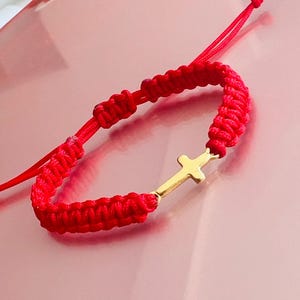 Può includere: Un braccialetto rosso intrecciato con un ciondolo a forma di croce color oro. Il braccialetto è realizzato con un cordoncino spesso e intrecciato e presenta una chiusura scorrevole regolabile. La croce è un design semplice ed elegante.
