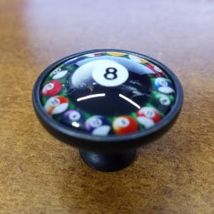 May include: A black cabinet knob with a glass top featuring a design of a pool ball. The center shows the number 8, surrounded by other pool balls in a green background. The knob is suitable for furniture.
