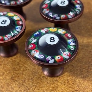 May include: A set of round cabinet knobs with a copper-toned metal base. The top of each knob features a glossy, domed graphic of a black eight ball surrounded by a ring of colorful numbered billiard balls, including numbers 2, 3, 4, 5, 7, 10, 11, 12, 13, 14, and 15.