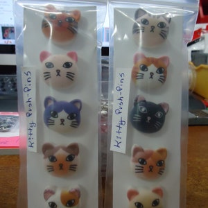 May include: Two sets of colorful cat-shaped push pins. Each set contains five pins with different cat designs. The pins are packaged in clear plastic bags with the text "Kitty Push-Pins" written on them. The text "SET #1" and "SET #2" is written on the top of each bag.