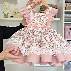 May include: A sleeveless dress featuring a white fabric with a pink rose and green leaf floral print. The dress has ruffled shoulders, two small pink bows at the waist, and a wide white lace trim along the hemline over a solid pink skirt.