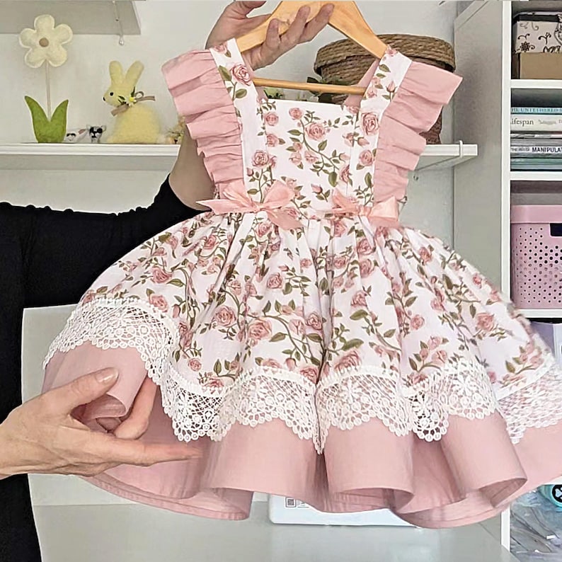 May include: A sleeveless dress featuring a white fabric with a pink rose and green leaf floral print. The dress has ruffled shoulders, two small pink bows at the waist, and a wide white lace trim along the hemline over a solid pink skirt.
