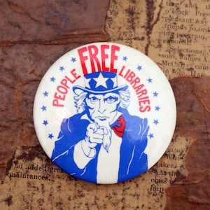 Puede incluir: Un botón blanco con una ilustración azul y roja del Tío Sam apuntando con el texto "PEOPLE'S FREE LIBRARIES" alrededor de la imagen.