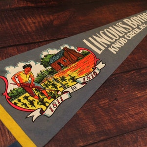 May include: A vintage pennant with a blue background and yellow trim. It features a depiction of a log cabin and a man working in a field of corn. The text reads "LINCOLN'S BOYHOOD HOME KNOB CREEK, KY. 1811 to 1816".
