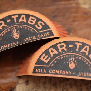 Puede incluir: Dos paquetes vintage de Ear-Tabs de color naranja y negro con el texto "U.S. Patent 2070332" y "Jola Company - Vista, Calif."