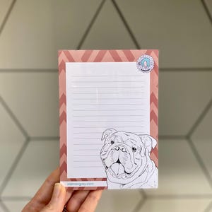 May include: A lined notepad featuring a bulldog illustration. The notepad has a pink and white chevron border and a small circular sticker. The website address oldmangrey.com is printed at the bottom.