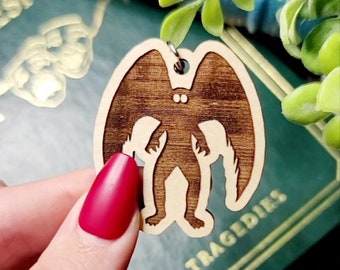 May include: A wooden keychain featuring a silhouette of a creature with wings. The keychain is brown and has a light wood grain. The creature has two large eyes and is standing with its wings spread out.
