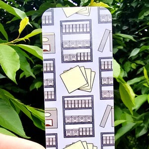 May include: A rectangular bookmark with a repeating pattern of bookshelves, books, and notepads. The background is a light blue-gray. The bookmark is held by a hand in front of a background of green leaves and grass.