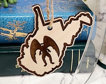 May include: Wooden ornament shaped like the state of West Virginia with a silhouette of a winged creature inside. The ornament is hanging from a twine string and is displayed on a clear acrylic stand.