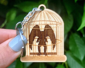 May include: Wooden keychain shaped like a birdcage with a silhouette of a bird with wings spread inside. The keychain is made of light brown wood and has a silver metal chain.