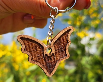 Pode incluir: Um chaveiro de madeira em forma de morcego com uma corrente e um anel prateados. O chaveiro tem a forma de um morcego com um design detalhado. O morcego é castanho e tem um contorno castanho claro. O chaveiro está pendurado numa mão sobre um fundo desfocado de flores amarelas e folhagem verde.