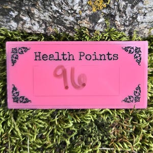 May include: A rectangular pink sign with black decorative corner flourishes and the text "Health Points" printed in black. The number "96" is written in pink marker in the center of the sign. The sign has a smooth, flat surface and edges.