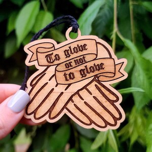 May include: Wooden ornament with a hand-drawn design of two hands clasped together. The ornament is brown and has a black string attached to it. The ornament has the text "To glove or not to glove" written on it.