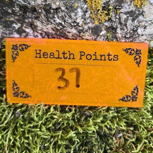 May include: A rectangular orange acrylic token featuring the black text Health Points and the number 37. The token has decorative black filigree corner accents and a subtle etched floral pattern across its surface, presenting a clear and legible design.