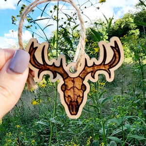 May include: A wooden deer skull ornament with a twine hanger. The ornament is brown and has a detailed design. The ornament is hanging in front of a field of green grass and yellow flowers.