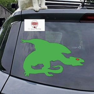 May include: A white and gray cat sits on the roof of a gray car. The car has a green dragon silhouette sticker on the back window. The sticker has a red eye. The car is parked in front of a wooden barn.