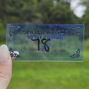 May include: A rectangular, translucent blue acrylic card featuring ornate black corner flourishes and the text "Health Points" printed in black. Below the text, the number "78" is written in a bold, dark marker on the smooth surface.