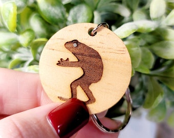 May include: A wooden keyring with a laser-cut design of a frog. The keyring is brown and has a silver metal ring.