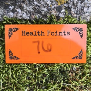 May include: A rectangular orange plastic sign featuring the black text "Health Points" at the top and the handwritten number "76" in the center. The sign is decorated with black filigree corner accents on all four sides of the rectangular border.