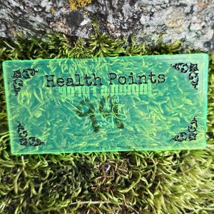 May include: A rectangular, translucent neon green acrylic token featuring black decorative corner flourishes and the text "Health Points" printed in a bold, black serif font across the center of the surface, with additional black text below.