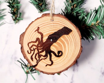 May include: A wooden Christmas ornament featuring a silhouette of an octopus, painted in black, on a natural wood slice. The ornament is hanging from a twine string.