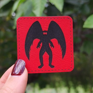 Op de afbeelding: Rode vierkante patch met een zwart silhouet van een wezen met vleugels en grote ogen. Het wezen heeft een humanoïde vorm met lange armen en benen.